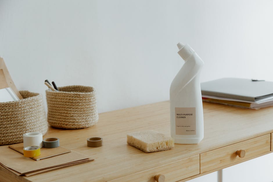 A wooden desk surface with a light natural finish, displaying a white multi-purpose cleaning spray bottle with a beige label, a yellow cleaning sponge, and several rolls of adhesive tape in white, gra