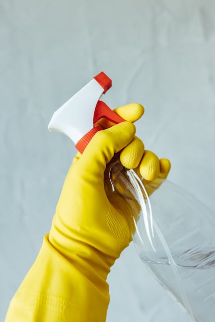 A person wearing bright yellow rubber gloves is holding a spray bottle filled with a cleaning solution, preparing for surface cleaning. The background features a plain, light-colored wall, highlightin