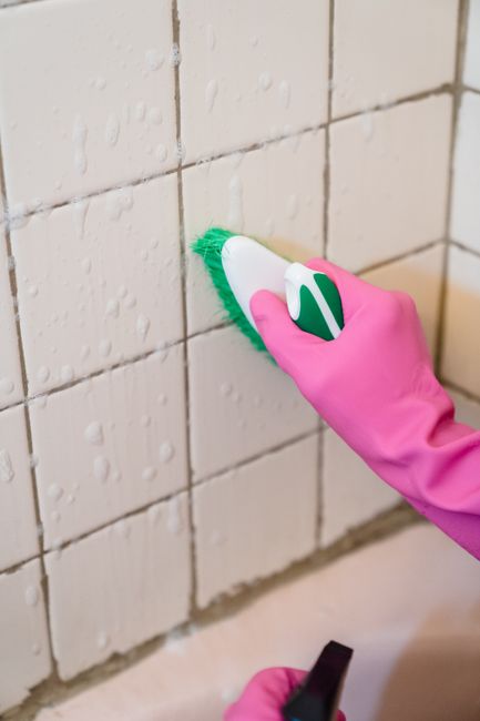 A person wearing bright yellow rubber gloves is holding a spray bottle filled with a cleaning solution, preparing for surface cleaning. The background features a plain, light-colored wall, highlightin