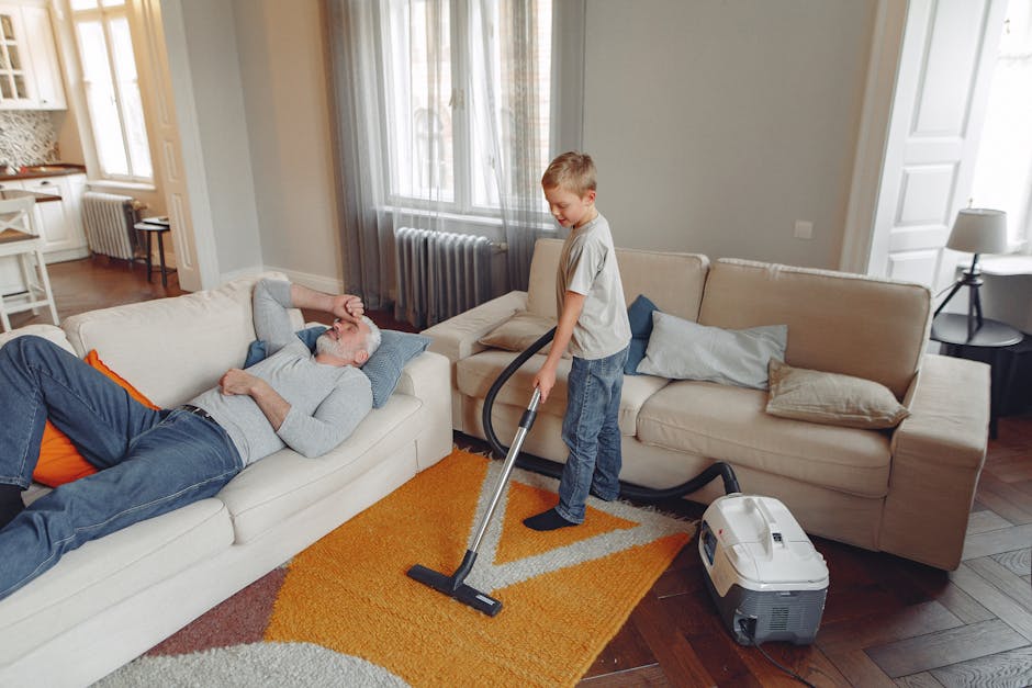 A young boy is vacuuming a beige fabric sofa in a well-lit living room, while an older man with gray hair and a beard reclines on the sofa, appearing relaxed. The room features wooden flooring partly 